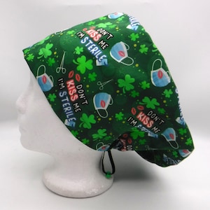 Könnte beinhalten: Grüne OP-Haube mit einem Muster aus Kleeblättern, Gesichtsmasken und dem Text "DON'T KISS ME I'M STERILE". Die Haube hat einen Kordelzug zur Anpassung und wird auf einem weißen Schaufensterkopf präsentiert.