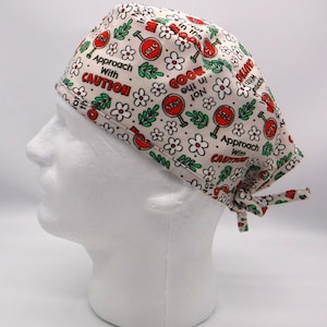Puede incluir: Un gorro quirúrgico de color crema con un patrón repetido de señales de stop rojas, flores blancas y hojas verdes. El gorro presenta las frases "Approach With Caution" y "Not in the Mood".