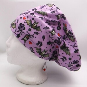 Puede incluir: Un gorro quirúrgico lavanda con un patrón repetido de murciélagos, flores y corazones. Los murciélagos tienen las alas extendidas y están en varias poses. El gorro está hecho de tela y tiene un cierre de cordón. El fondo es blanco.