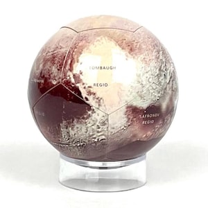 May include: A red and white globe model of Pluto with the names "Tombaugh Regio" and "Saronov Regio" printed on the surface. The globe is on a clear plastic stand.