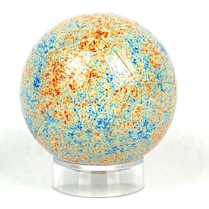 May include: A decorative sphere with a speckled pattern in blue, orange, and yellow, reminiscent of a cosmic map. The sphere is displayed on a clear stand, creating a floating effect. The overall theme is celestial and artistic. The sphere is approximately 15 cm in diameter.