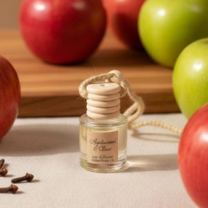 May include: A small glass car diffuser bottle with a light wood cap and a beige hanging cord. The label reads Applewood & Clove car diffuser limerluxe ca. The bottle is surrounded by several whole red and green apples and small brown cloves.
