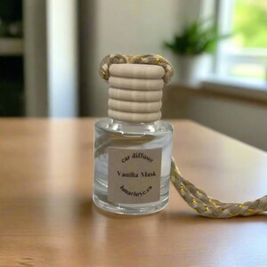 May include: A clear glass car diffuser with a wooden top and a braided rope. The label reads "car diffuser Vanilla Musk lumarluxe.ca". The wooden top has horizontal ridges. The rope is beige with gold accents.