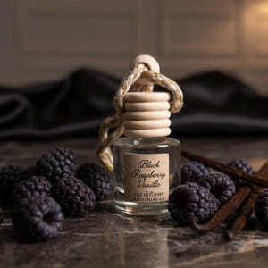 May include: A small, clear glass car air freshener bottle with a wooden cap and a rope. The label reads "Black Raspberry Vanilla". Dark raspberries and vanilla pods are arranged around the bottle. The background is dark.