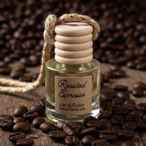 May include: A clear glass car air freshener bottle with a wooden top and a rope. The label reads "Roasted Espresso" and "car diffuser lunarluxe.ca". The bottle is surrounded by roasted coffee beans on a wooden surface.