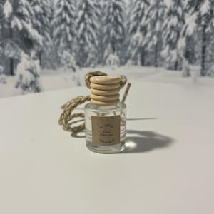 May include: A small, clear glass car air freshener with a wooden cap and a beige string. The label reads "White Christmas". The background features a snowy forest scene.