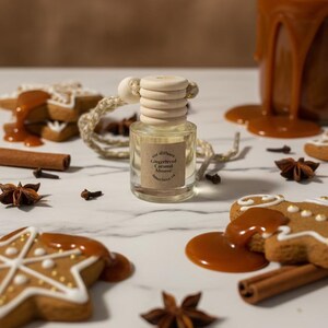 Peut inclure: Un flacon de parfum de voiture en verre transparent avec un bouchon en bois, étiqueté "Gingerbread Caramel Mousse". Des biscuits en pain d'épices avec du glaçage blanc et de la sauce au caramel sont dispersés autour du flacon, avec des bâtons de cannelle et de la badiane.