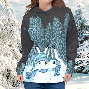Peut inclure: Un sweat-shirt noir avec une scène d'hiver représentant deux lapins blancs portant des écharpes et des bonnets bleus. Les lapins se tiennent dans une forêt enneigée avec des sapins bleus.