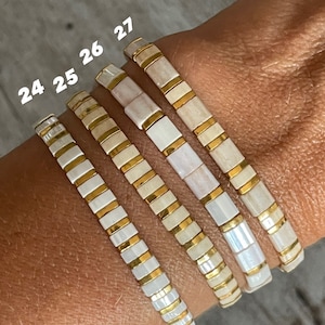 May include: Four white and gold bracelets stacked on a wrist. The bracelets are made of small square tiles with a white background and gold trim. The numbers 24, 25, 26, and 27 are written on the bracelets.