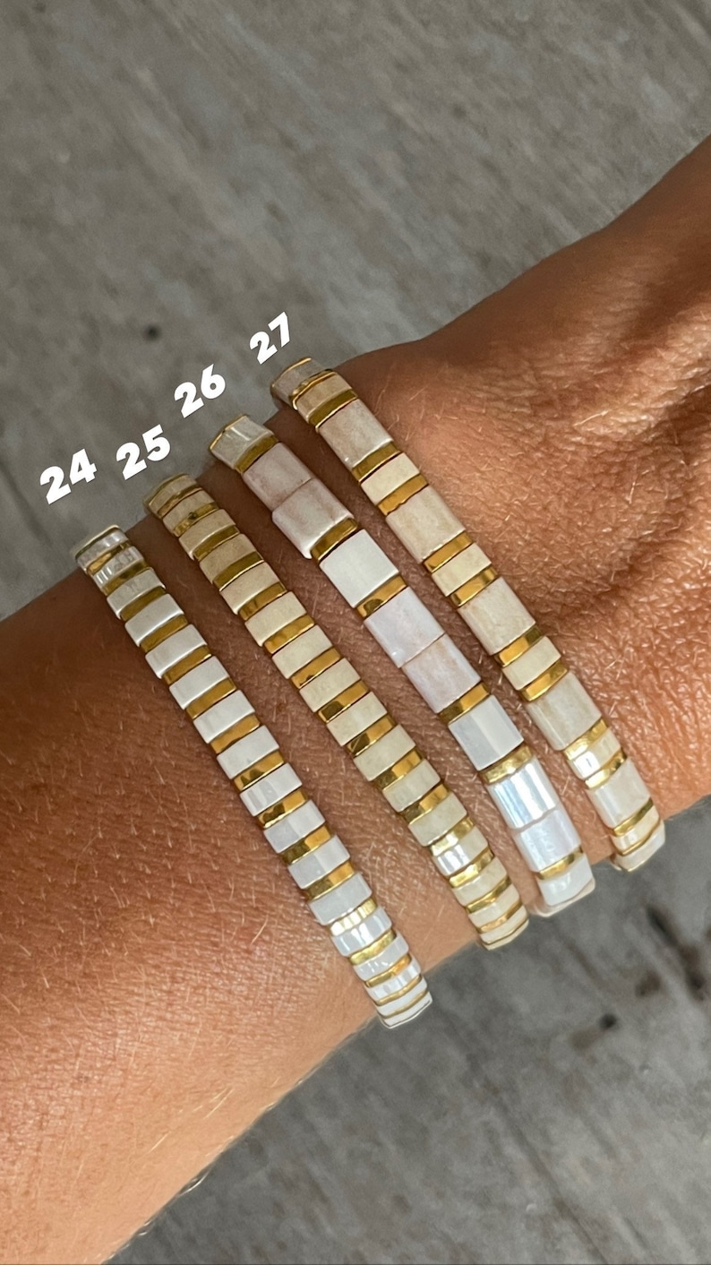 May include: Four white and gold bracelets stacked on a wrist. The bracelets are made of small square tiles with a white background and gold trim. The numbers 24, 25, 26, and 27 are written on the bracelets.