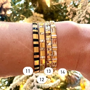 May include: Four gold and white tile bracelets stacked on a wrist. The bracelets are numbered 11, 12, 13, and 14.