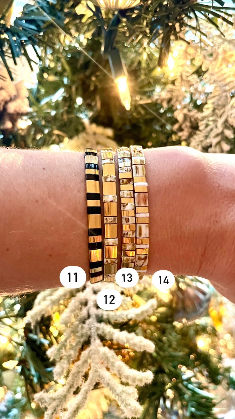 May include: Four gold and white tile bracelets stacked on a wrist. The bracelets are numbered 11, 12, 13, and 14.