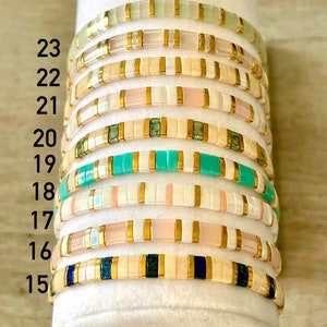 May include: A stack of 9 colorful beaded bracelets with gold accents. The bracelets are numbered 15 through 23.