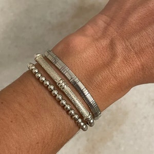 Puede incluir: Tres pulseras de plata apiladas en la muñeca de una persona. La pulsera superior es una banda de plata con un diseño texturizado. La pulsera del medio es una banda de plata con un diseño trenzado. La pulsera inferior es una pulsera de plata con cuentas.