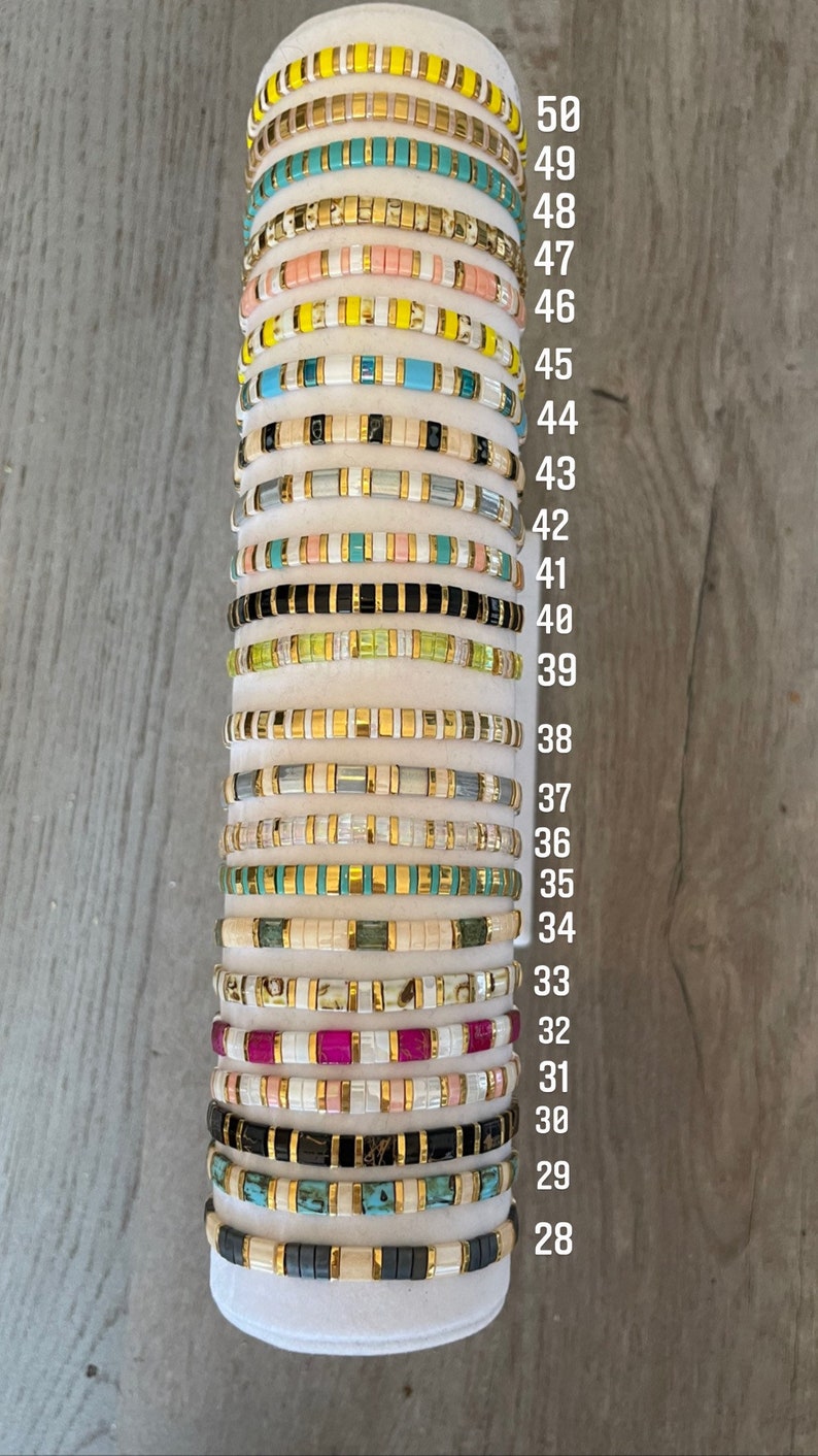 May include: A stack of 23 colorful beaded bracelets with gold accents. The bracelets are numbered from 28 to 50.