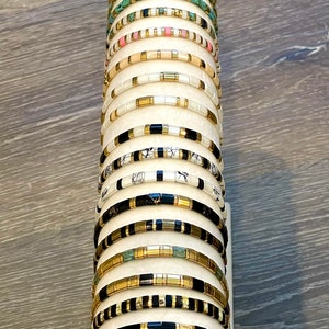 May include: A stack of colorful beaded bracelets on a white display stand. The bracelets are made of various materials, including gold, black, white, and turquoise.