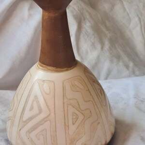 Puede incluir: Un jarrón de cerámica blanca con cuello y base marrones. El jarrón tiene un patrón geométrico de líneas blancas sobre un fondo blanco.