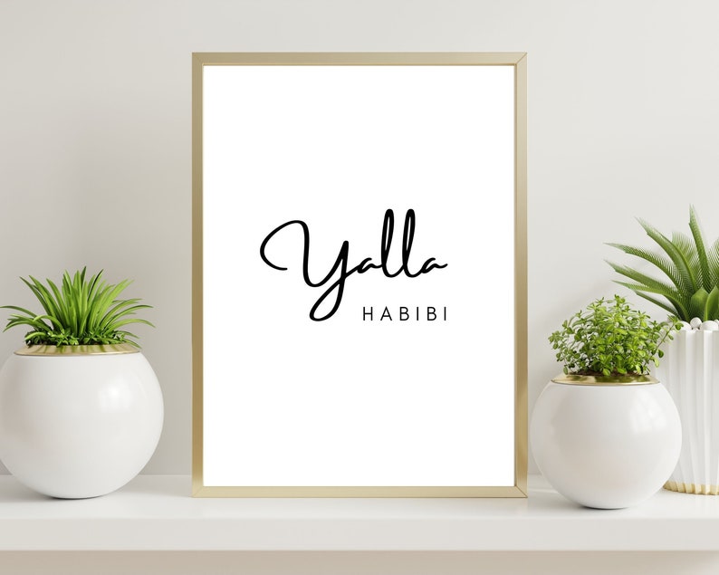 May include: A framed print with the words "Yalla Habibi" in black script on a white background. The frame is gold. The print is displayed on a white shelf with potted plants on either side.