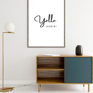 May include: A framed white print with the words "Yalla Habibi" in black script. The print hangs above a wooden cabinet with a teal blue sliding door. A gold floor lamp and a black vase are also visible.