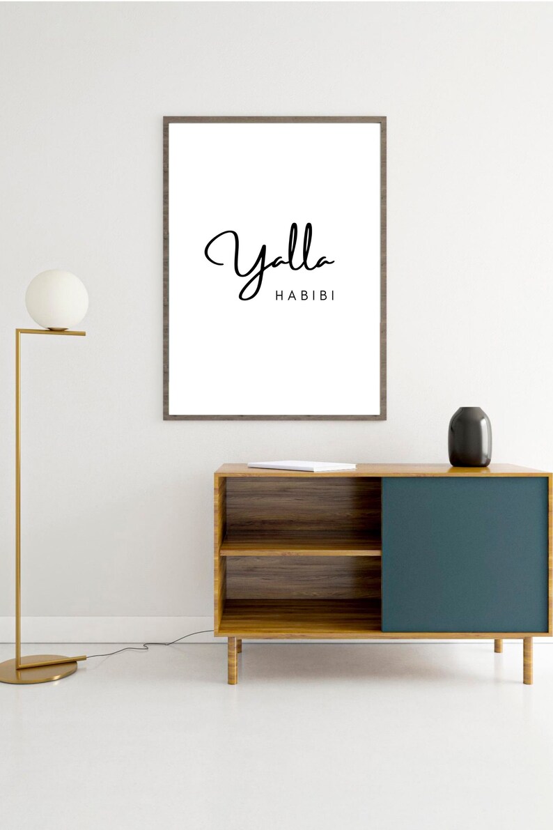 May include: A framed white print with the words "Yalla Habibi" in black script. The print hangs above a wooden cabinet with a teal blue sliding door. A gold floor lamp and a black vase are also visible.