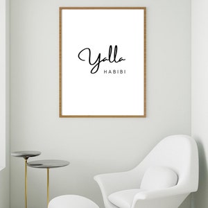 May include: A framed art print with the words "Yalla Habibi" in black script on a white background. The frame is a light brown color. The print is displayed in a modern living room setting with a white chair and side tables.