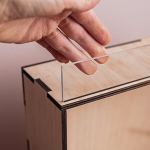 May include: A hand places a clear acrylic panel into a wooden box with a black trim. The box is made of light wood and has a natural finish.