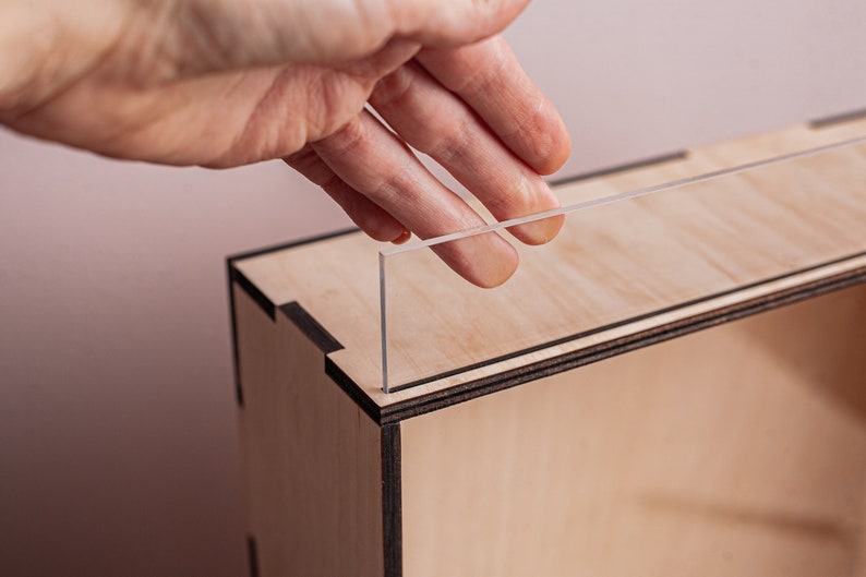 May include: A hand places a clear acrylic panel into a wooden box with a black trim. The box is made of light wood and has a natural finish.