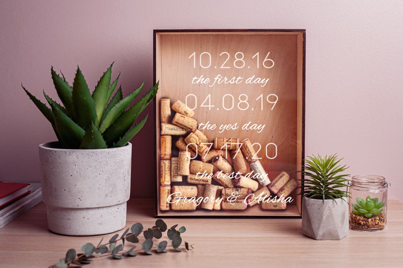 May include: A wooden shadow box with a glass front filled with cork wine bottle stoppers. The box has a white inscription that reads "10.28.16 the first day 04.08.19 the yes day 07.17.20 the best day Gragory & Alisha".
