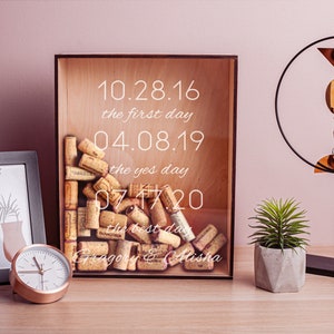 May include: A wooden box with a glass front filled with wine corks. The box has the dates 10.28.16, 04.08.19, and 07.17.20 printed on it, along with the text "the first day", "the yes day", and "the best day". The box also has the names "Gregory & Alisha" printed on it.