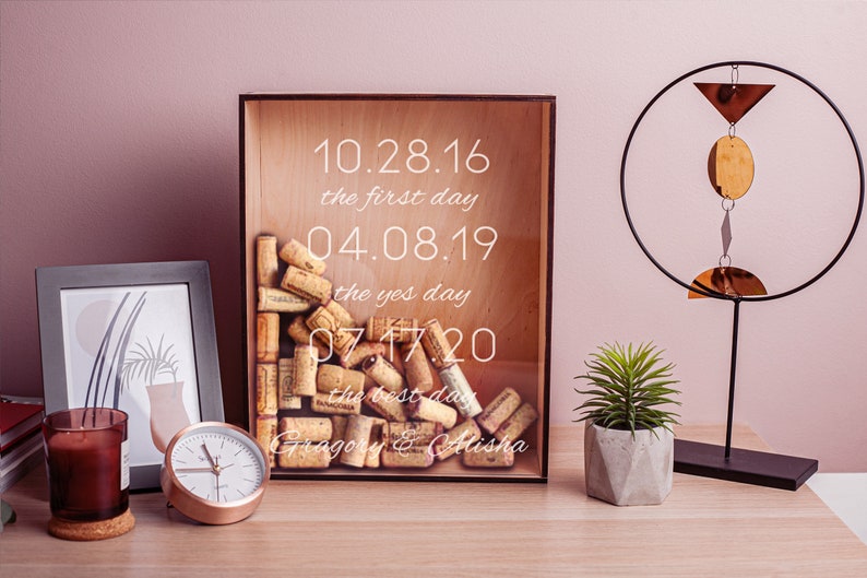 May include: A wooden box with a glass front filled with wine corks. The box has the dates 10.28.16, 04.08.19, and 07.17.20 printed on it, along with the text "the first day", "the yes day", and "the best day". The box also has the names "Gregory & Alisha" printed on it.