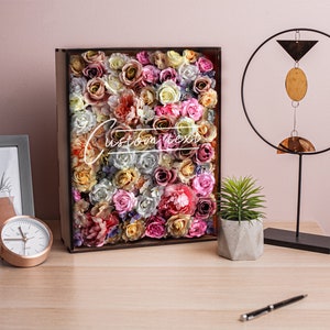 May include: A wooden box filled with a variety of colorful artificial flowers, including roses, peonies, and carnations. The box is on a wooden table with a clock, a candle, and a small potted plant.