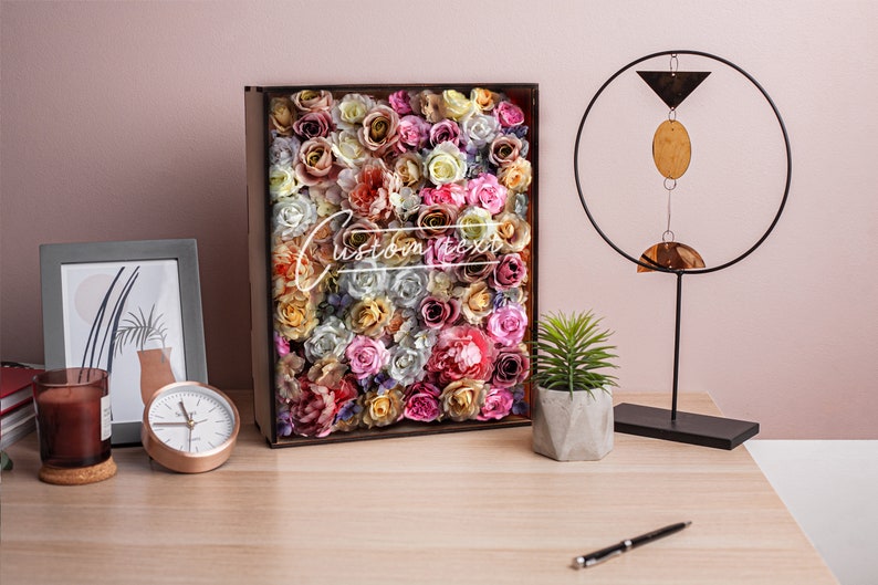 May include: A wooden box filled with a variety of colorful artificial flowers, including roses, peonies, and carnations. The box is on a wooden table with a clock, a candle, and a small potted plant.