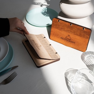 May include: Two wooden menu holders with a brown wood finish and the text "Restaurant" engraved on the front. The menu holders are open and have a light wood interior.