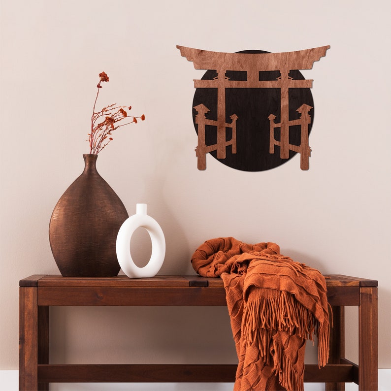 May include: A wooden wall decor featuring a Japanese torii gate with a black circle behind it. The gate is brown and the circle is black. The decor is mounted on a light beige wall. A brown vase and a white vase are on a wooden table in front of the decor. A brown and orange throw blanket is draped over the table.