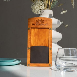 May include: A wooden restaurant menu holder with a light brown finish. The top features a logo and the text "RESTAURANT LOGO". The holder has a black section at the bottom and is secured with brass-coloured rivets. A clear glass and blue plates are in the background.