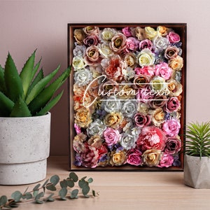 May include: A wooden frame filled with a variety of artificial flowers in shades of pink, white, and yellow. The flowers are arranged in a dense pattern, creating a colorful and textured display. The frame is sitting on a wooden table, next to a potted succulent and a glass jar with a small succulent.