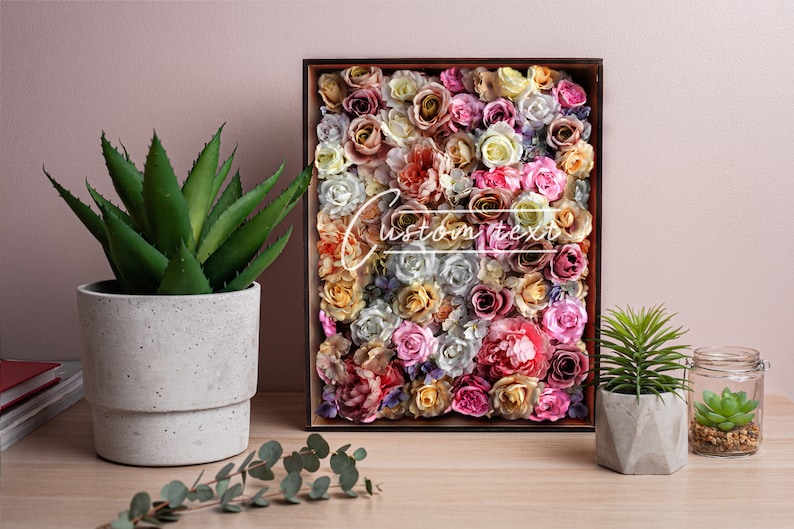 May include: A wooden frame filled with a variety of artificial flowers in shades of pink, white, and yellow. The flowers are arranged in a dense pattern, creating a colorful and textured display. The frame is sitting on a wooden table, next to a potted succulent and a glass jar with a small succulent.