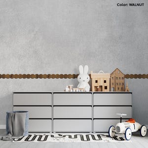 Può includere: Una moderna scena di cameretta con una cassettiera grigio chiaro a sei cassetti. La cassettiera è adornata con casette giocattolo in legno, un coniglietto bianco e un'auto giocattolo. Un cesto blu con una coperta grigia e un tappeto con motivo stradale completano l'arredamento.