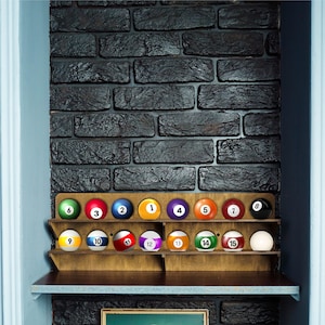 May include: A wooden rack with 15 billiard balls, numbered 1 through 15, in a variety of colors. The rack is on a shelf against a brick wall.