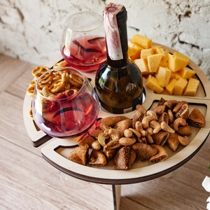Op de afbeelding: Een houten serveerblad met vier compartimenten voor snacks en drankjes. Het blad heeft een ingebouwde flessenhouder en twee glazenhouders. Het blad is gevuld met snacks, waaronder pretzels, vijgen, cashewnoten en kaas.
