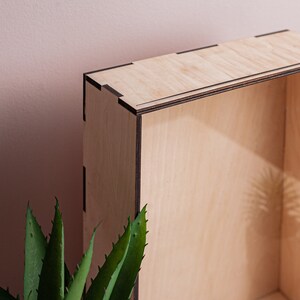 May include: A wooden box with a natural finish and black trim. The box is empty and has a shadow of a plant inside.