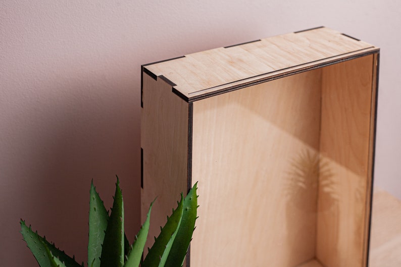May include: A wooden box with a natural finish and black trim. The box is empty and has a shadow of a plant inside.