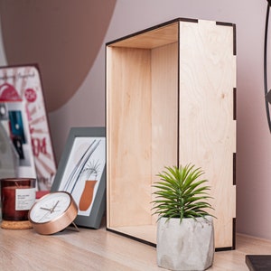 May include: A wooden box with a black trim sits on a wooden table. A small potted plant sits inside the box. A clock, a candle, and a picture frame are on the table in front of the box.