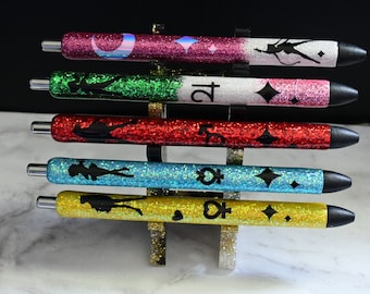 May include: Five glitter pens with black silhouettes of a person, various symbols, and glitter accents. The pens are arranged vertically in a stand and are in the colors pink, green, red, blue, and yellow.