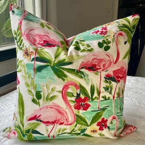 May include: Decorative pillow with a tropical flamingo design. The pillow features pink flamingos, red flowers, and green foliage on a white background. The pillow is square-shaped and appears to be made of fabric.