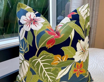 May include: Square throw pillow with a tropical floral pattern. The pillow features large white hibiscus flowers with red centers, orange and red flowers, and green leaves against a navy blue background. The pillow is on a white quilted bedspread.