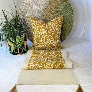 May include: A gold and white leaf patterned table runner with tassels on each end. The runner is folded over a matching pillow. The pillow and runner are on a white surface.