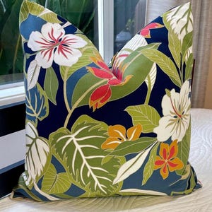 May include: Decorative throw pillow with a vibrant tropical floral pattern. The pillow features large white hibiscus flowers with red centers, orange and red blooms, and green leaves against a navy blue background. The pillow is placed on a white bedspread.