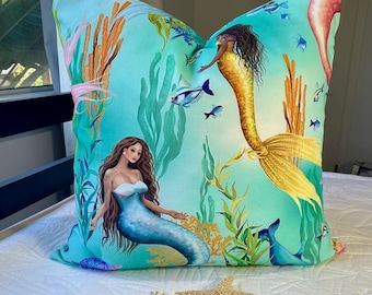 May include: A turquoise blue pillow with a mermaid design. The pillow features two mermaids with long flowing hair, one with a blue tail and the other with a yellow tail. The mermaids are surrounded by seaweed and coral.