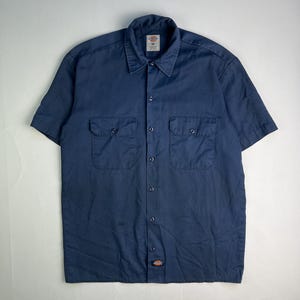 May include: Navy blue short-sleeve button-up shirt with two chest pockets. The shirt has a collar and a small logo patch near the bottom. The shirt is a medium size.